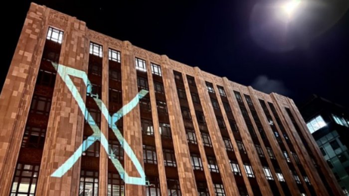 A nighttime view of a large brick building with a modern 'X' logo projected onto its facade, illuminated under a bright moonlight.