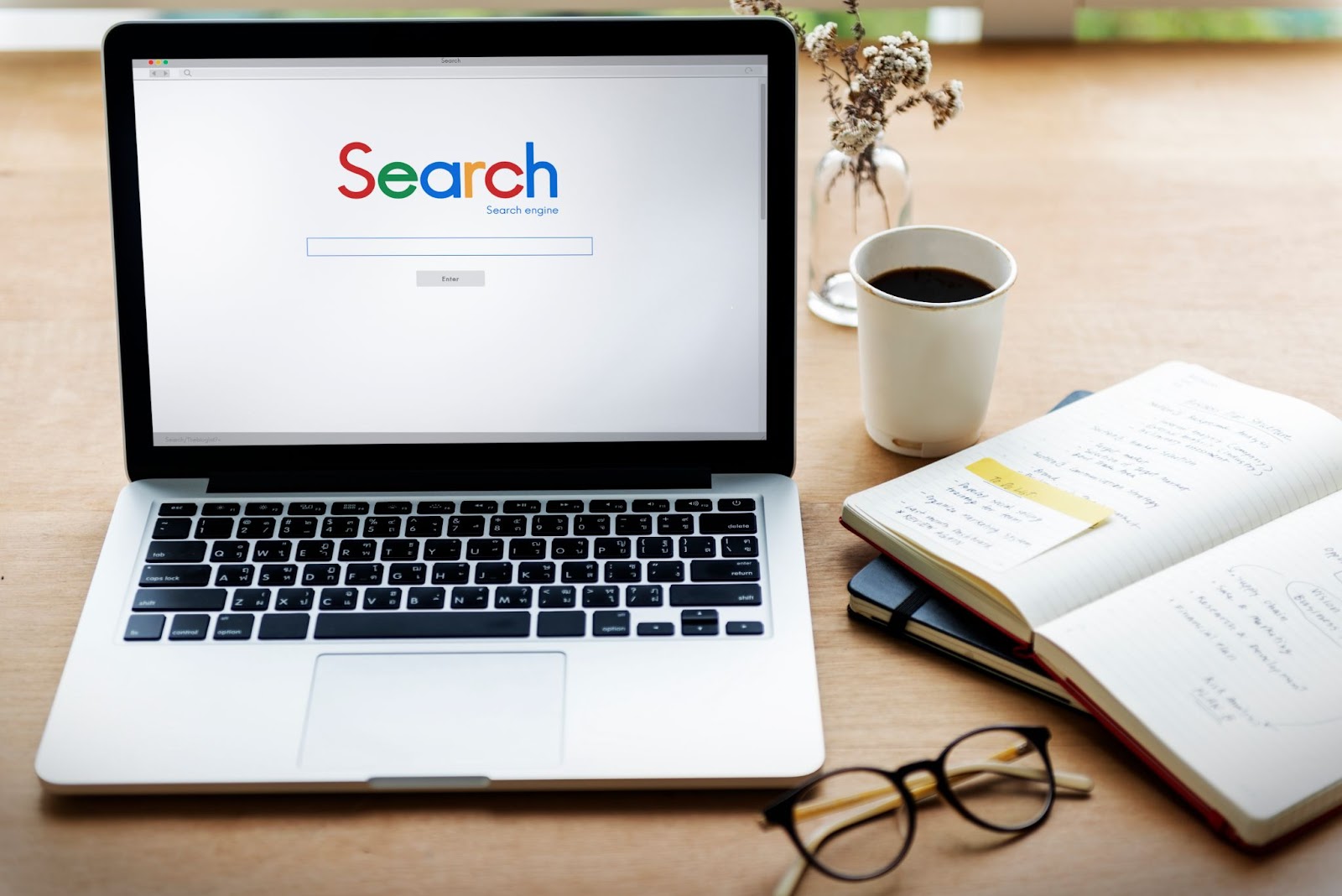 Laptop displaying a search engine on the screen, next to a notebook, glasses, and a cup of coffee on a desk.