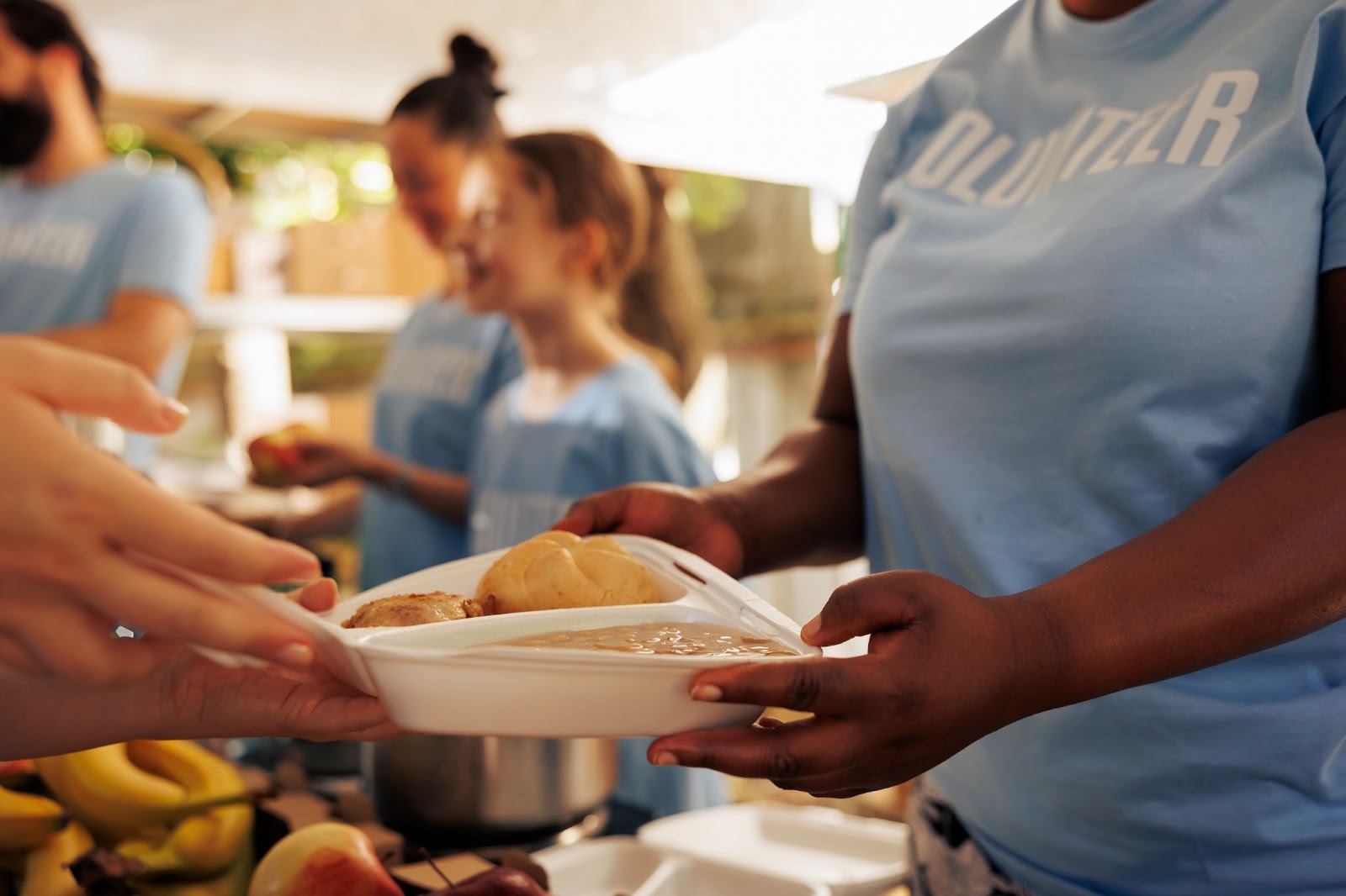Volunteer handing a plate of food to a person, with other volunteers and fruits in the background during a community event.
