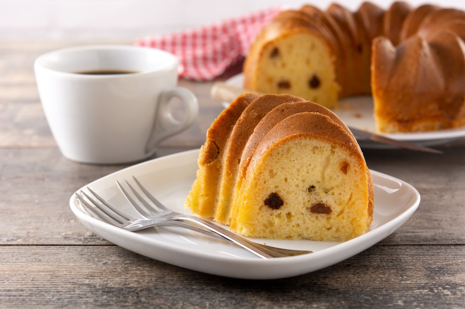 Slice of bundt cake with raisins served on a plate with two forks, accompanied by a cup of coffee.